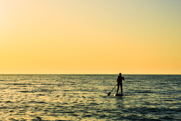 Fototapeta premium The silhouette of a man on a board stands and holds an oar at sea