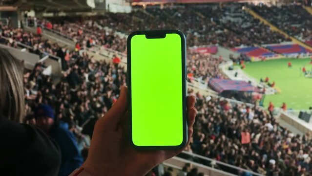 A woman watches football at the stadium, holds a phone with a green screen in hands. Makes bets on sports.