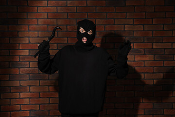 Emotional thief in balaclava raising hands with crowbar against red brick wall