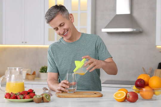 Smiling man pouring fresh orange juice into glass at white marble table in kitchen - Powered by Adobe