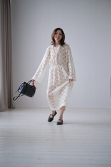 woman in a white lace dress stands gracefully in a minimalistic room, holding a black handbag. Her...