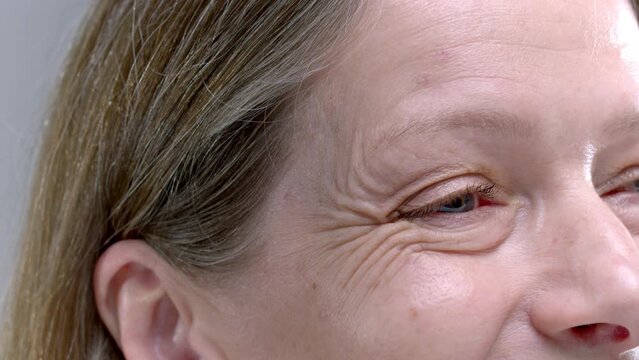 Happy senior woman with blue eyes and deep crows feet laughs on light grey background closeup. Old female person expresses real happiness and joy. Aged lady face with pampered wrinkled skin