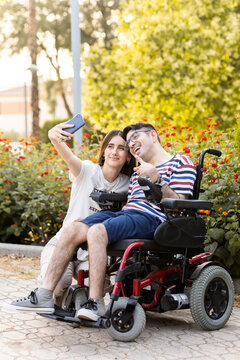 A young woman cares for her disabled brother in an automatic wheelchair at an outdoor park.The disabled woman and man take a selfie with the mobile phone. Concept of men with cerebral palsy.