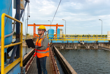 an engineer stand beside the blue water tank and point his finger forward while holding laptop on his left hand and checking the switch behind the water tank at waste water treatment