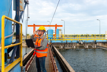 Fototapeta premium an engineer stand beside the blue water tank and his right hand touch his glasses while holding laptop on his left hand and checking the switch behind the water tank at waste water treatment