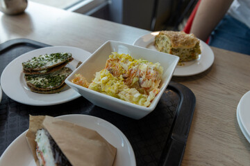 A close-up shot of a lunch table featuring a variety of delicious dishes, including a creamy salad, a savory pastry, and a wrapped sandwich. The bright, natural light suggests a sunny day