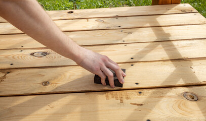 A close-up shot of a hand sanding a wooden deck board with a sanding block. The natural light...