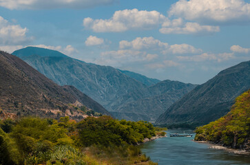 Photograph of a canyon during the summer season with a blue sky with some clouds, two mountains with a lot of nature and a river in the middle of them
