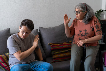An adult son and his mother raise their hands in frustration during a heated argument while sitting...