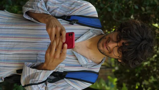 A young man with curly short hair and a light shirt smiles while texting on his smartphone in a vertical video, conveying a cheerful and engaged demeanor