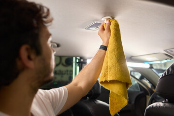 A man is cleaning the cars ceiling meticulously with a yellow cloth, focusing on detail