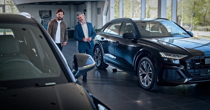 Cheerful man is talking to dealer with car keys. Another car dealer is passing by. Bearded caucasian man dealer shows customer car model and specification of the car