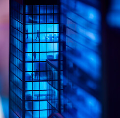 A modern glass office building with blue lights inside at night. Grid-like pattern of windows visible, with small moving figures inside.