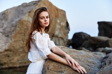 Serene young woman in white dress sitting on rocky shore of tranquil water