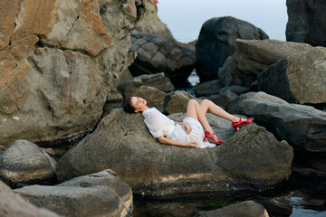 serene asian woman in white dress relaxing on rocky ocean shore with vibrant red shoes