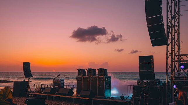 A wide shot of a concert stage setup on a beach at sunset. The stage is equipped with large speakers and lighting equipment, and the ocean can be seen in the background.