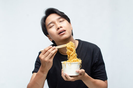 young Asian man enjoys his noodles with deep pleasure using chopstick and paper bowl. man in black tshirt eats noodles with great enthusiasm.