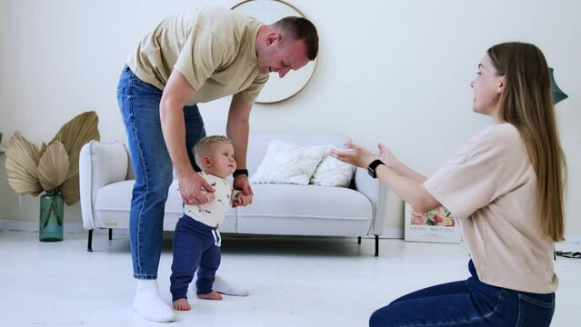 Father supports his cute little son as he learns to walk. Mom sits in front clapping and calling him up. Light living-room backdrop.