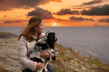 Portrait of a woman and an English Cocker Spaniel puppy.She sits on the rocky shore of the island and holds a dog in her hands.Both look into the distance.In the background there is a lake and sunset.