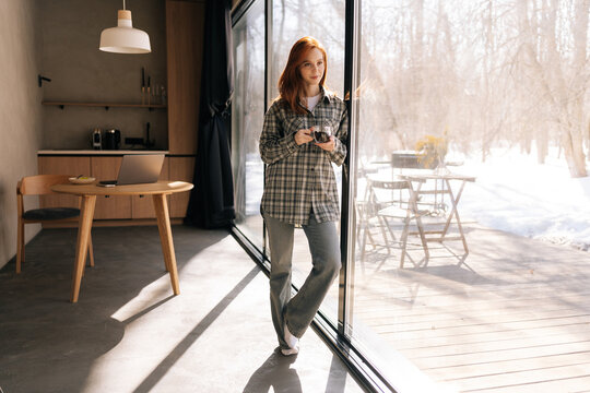 Full length portrait of pretty woman drinking enjoying hot coffee, smiling looking away, standing by window with snowy winter nature view on sunny morning. Happy redhead female having rest at home - Powered by Adobe