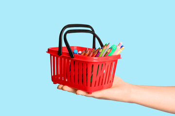 Female hand with red shopping basket and different school stationery on blue background