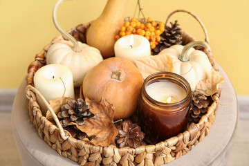 Wicker tray with burning candles, pumpkins, pine cones and autumn leaves on pouf near yellow wall