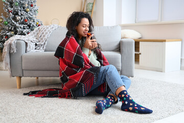 Beautiful young African-American woman with glass cup of hot mulled wine on carpet at home