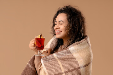 Beautiful young African-American woman with glass cup of hot mulled wine and plaid on brown...