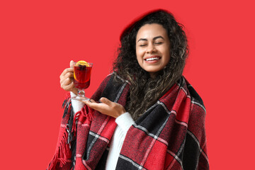 Beautiful young African-American woman with glass cup of hot mulled wine and plaid on red background