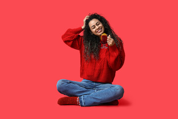 Happy young African-American woman with glass cup of hot mulled wine on red background