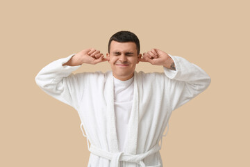 Portrait of handsome young man in bathrobe suffering from loud noise on beige background