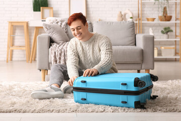 Happy young man with stylish suitcase sitting on floor at home