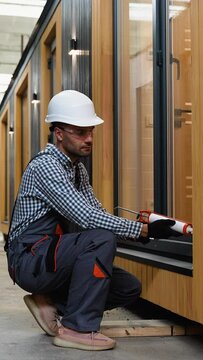 A worker is applying polyurethane foam to fill gap between sash and window frame in prefabricated modular house. Vertical video
