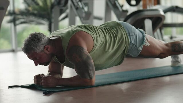 A muscular man performs a plank exercise on a yoga mat in a gym, showcasing strength and fitness dedication.