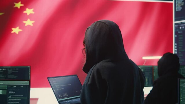 Chinese hacker working in governmental hacking room with Chinese flag on a big screen. Having the mission on inflicting fear and misinformation among the population. Dictatorship propaganda. Camera B.