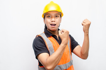 ecstatic asian male construction worker in yellow hardhat and orange vest raising his fist showing his success and celebrating his achievments.