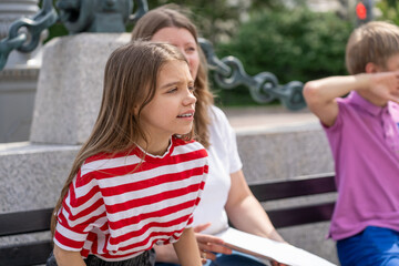 Young girl wearing red and white striped shirt sits on bench in park with her family. She looks to...