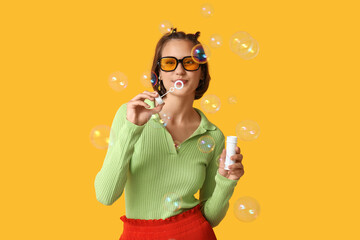 Teenage girl in sunglasses blowing soap bubbles on yellow background