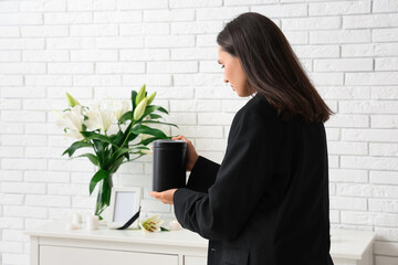 Young woman putting mortuary urn on chest of drawers near bouquet of funeral lily flowers in room