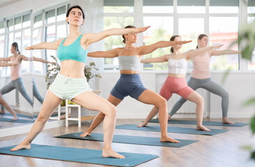 Obraz premium Girls during group hatha yoga workout. Female students perform maintain warrior pose, virabhadrasana, lightweight option for beginners and amateurs. Coach strike pose virabhadrasana