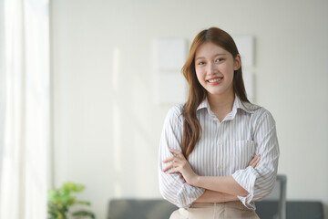 Confident woman with crossed arms, standing in bright room, wearing striped shirt, smiling at camera. Professional, business, indoor portrait.