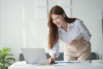 Smiling woman working on laptop at desk, holding tablet, modern office, business environment, professional setting, white interior decor.