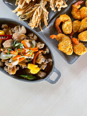 Sweet and sour sauce mushrooms with chopped pepper and pineapple on a hot plate, battered fried prawns and crispy enoki mushrooms on the table background. Copy space.