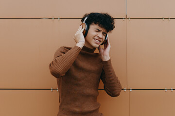 Young asian man in fashionable attire listening to music with headphones against a brown wall