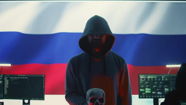 Digital outlaw hacker in a dark hood issuing cyber threats with a human skull to make death threats, aggressive malicious intent. Russian man committing cybercrime and scamming nations. Camera B.