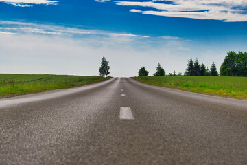 Empty asphalt road leading into horizon under blue sky with clouds and green fields