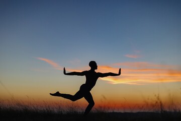 Silhouette of male ballet dancer doing a majestic dance move in nature, Behind him are heavenly clouds and the sun is setting.