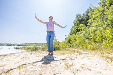 A woman happily dancing by the lake beach, enjoying the sunny day surrounded by greenery, carefree and joyful in nature