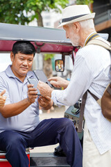 American senior tourist man poses paying the fare to the Tuktuk Thailand taxi driver by smart phone on blurred of city background. Senior tourist concept
