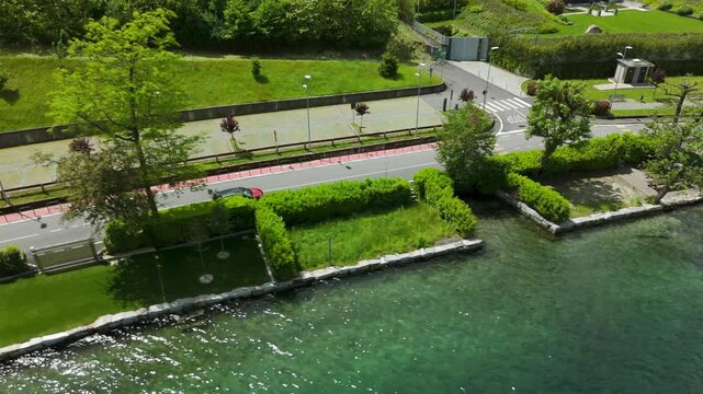 Tracking aerial view of red car on asphalt road next to lake in town, Pella, Italy